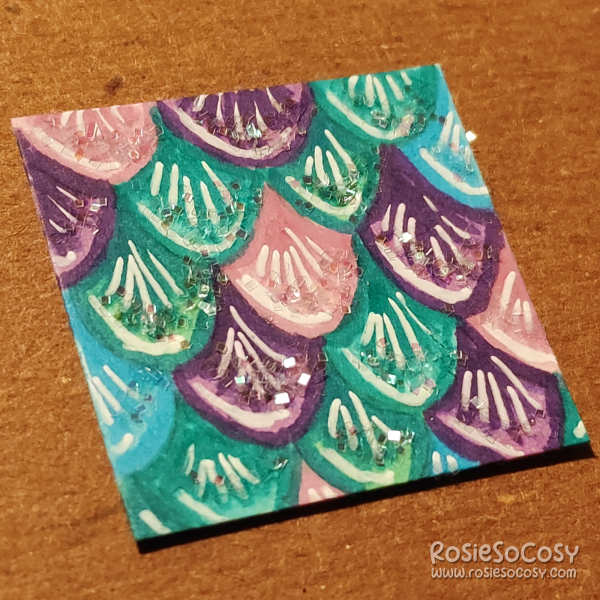 An inch sized drawing of teal, blue, purple and pink mermaid scales. There's some glitter on them.