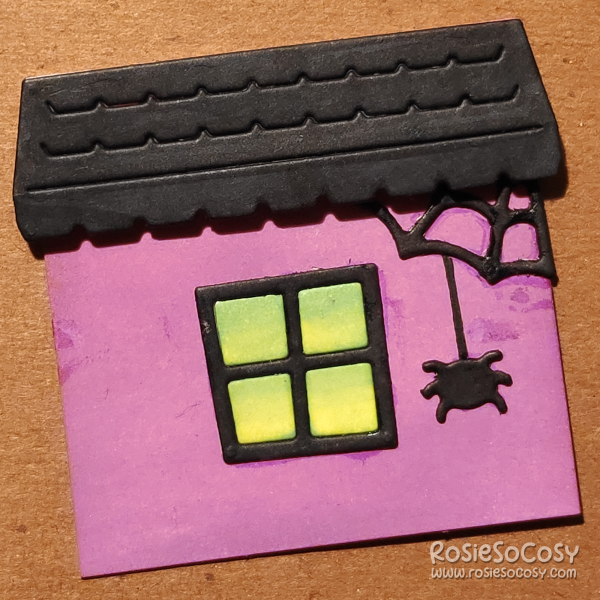 A two inch sized card consisting of a purple house, black window with green light coming from within the building, a black roof and a black spider hanging from a spiderweb in the top right corner.