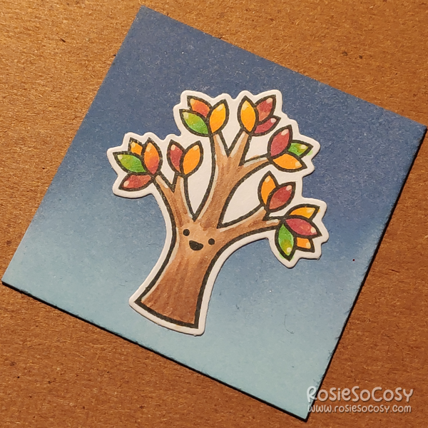 A tiny 2 inch card with a colourful tree. The tree is a medium brown colour, with a happy face, the leaves are green, yellow, orange and red. The background is a (dark to light) blue gradient.