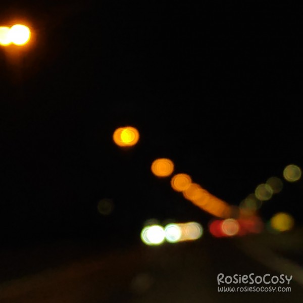 Driving on the highway. It's dark outside. Orange, red, yellow and white lights can be seen in the distance, with a bokeh effect.