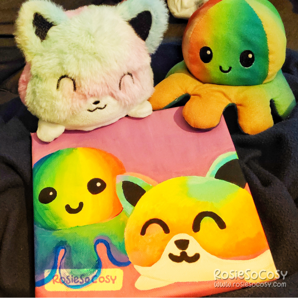 A canvas painting done in Posca markers, with two plushies on it. One is a rainbow octopus, the other a rainbow cat.