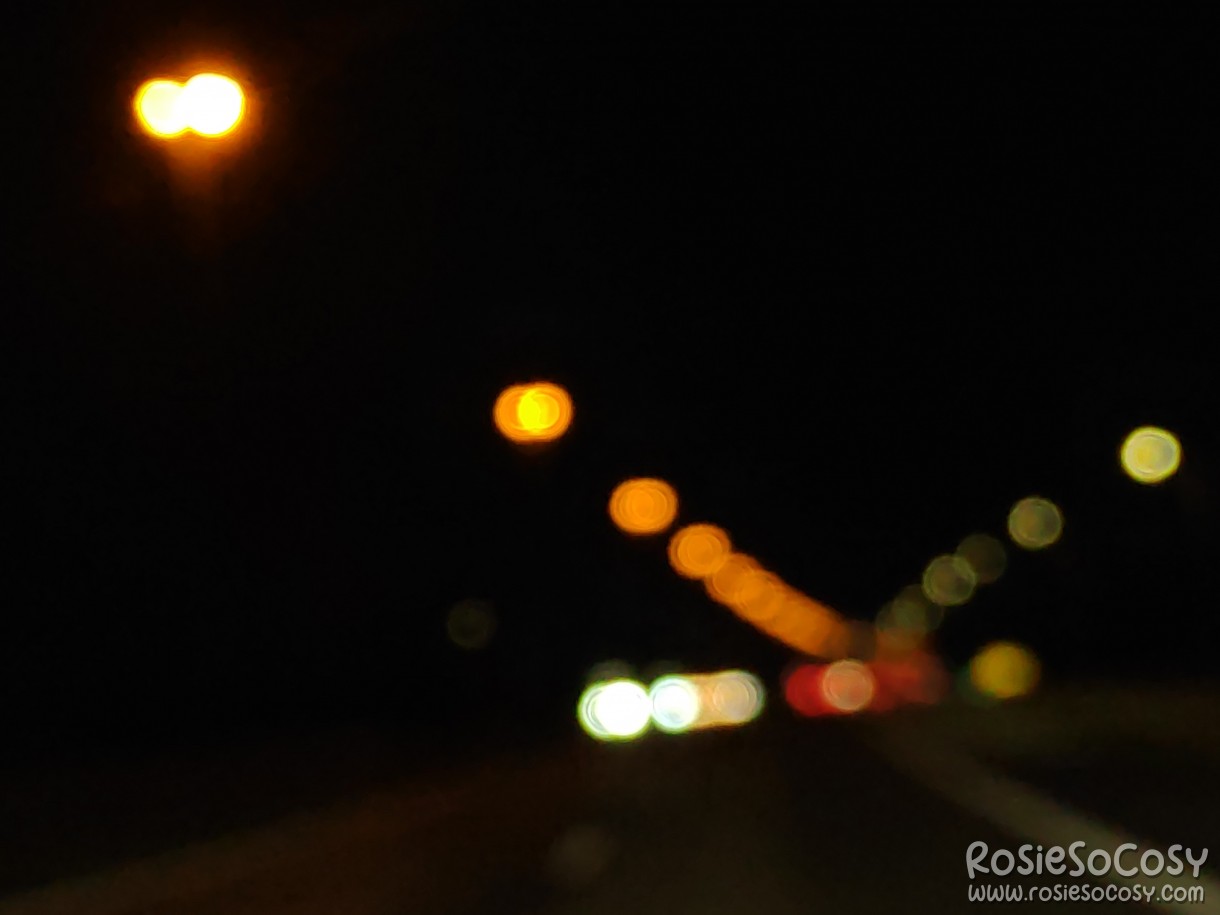 Driving on the highway. It's dark outside. Orange, red, yellow and white lights can be seen in the distance, with a bokeh effect.