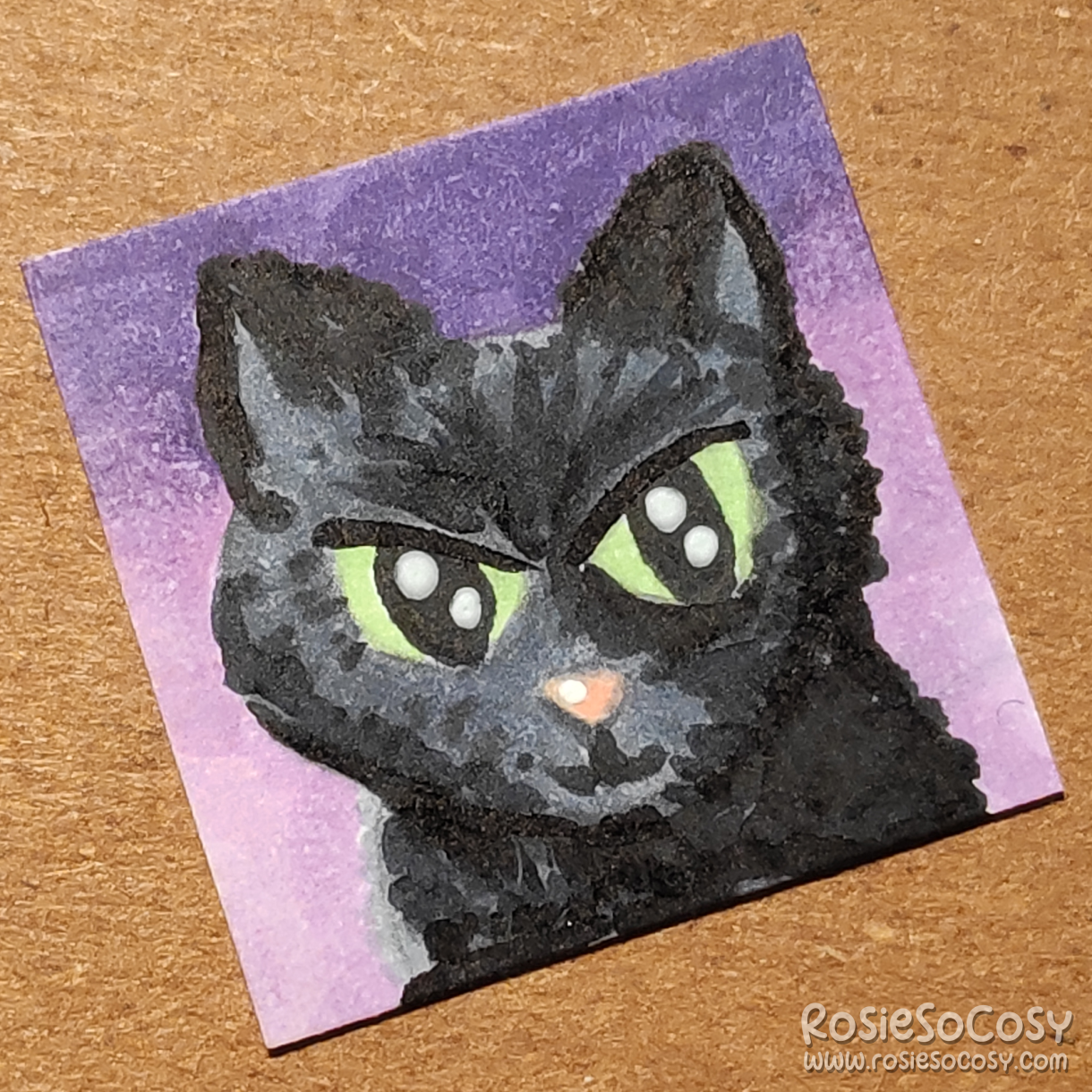 An inch sized drawing of a black cat with green eyes. The background is a purple and lilac gradient.