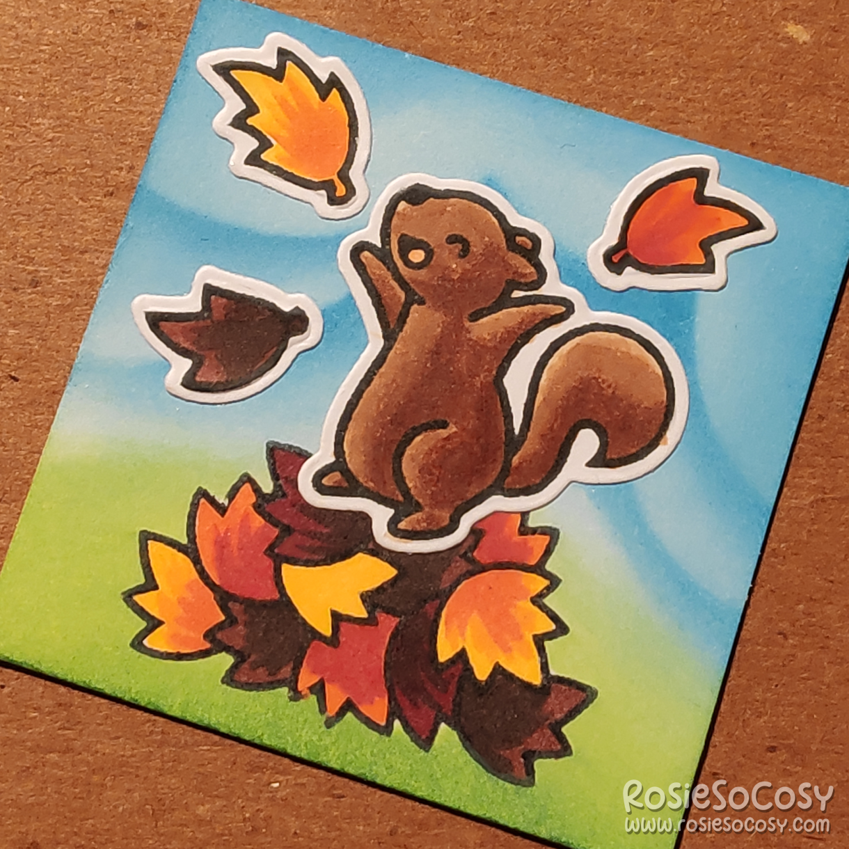 A tiny 2 inch card with a blue cloudy background, and a green grassy ground below, there is a overtly happy brown squirrel dancing atop a big pile of leaves, with a few of those leaves flying around.