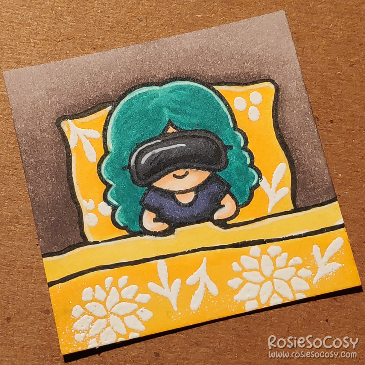 A tiny 2 inch card with a girl sleeping in a bed. She is wearing a black sleeping mask, a very dark bluish grey shirt, and her duvet is a bright yellow with white floral elements.