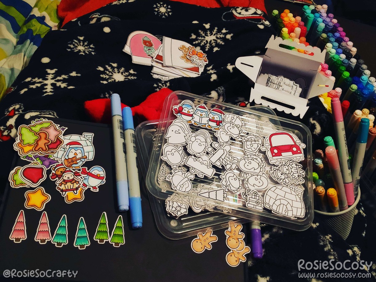 Rosie is sitting on the sofa, with craft supplies scattered everywhere. There are blankets on her legs, hundreds of stamped and cut images on lids, waiting to be coloured. Some images have already been coloured in with Copic markers. The images are all in a winter and Christmas theme; penguins, pine trees, Igloos, gingerbread cookies, snow globes.