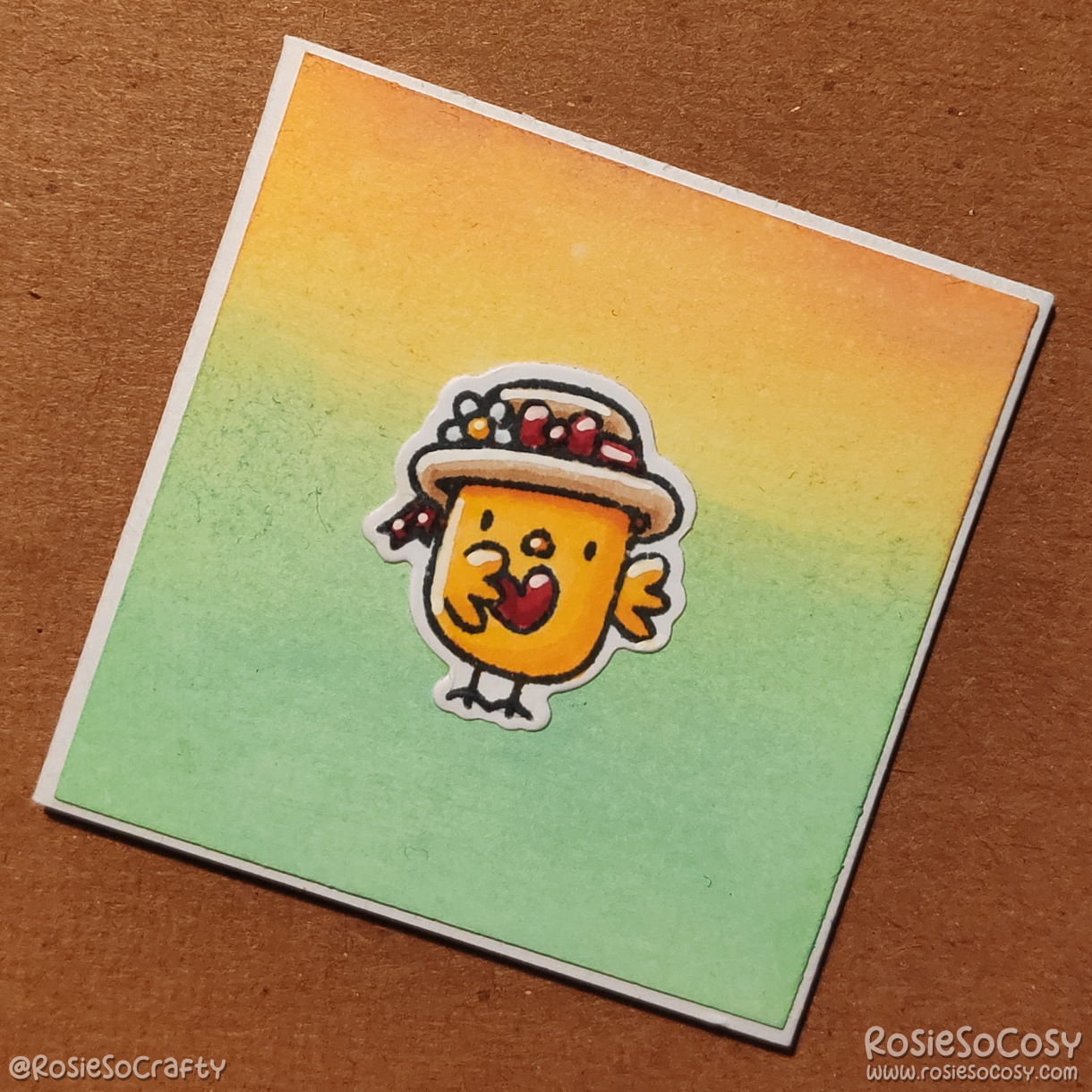 A inch sized card with a yellow to green gradient background. On the card is a tiny yellow chick with a cute floral hat, holding a tiny red heart.