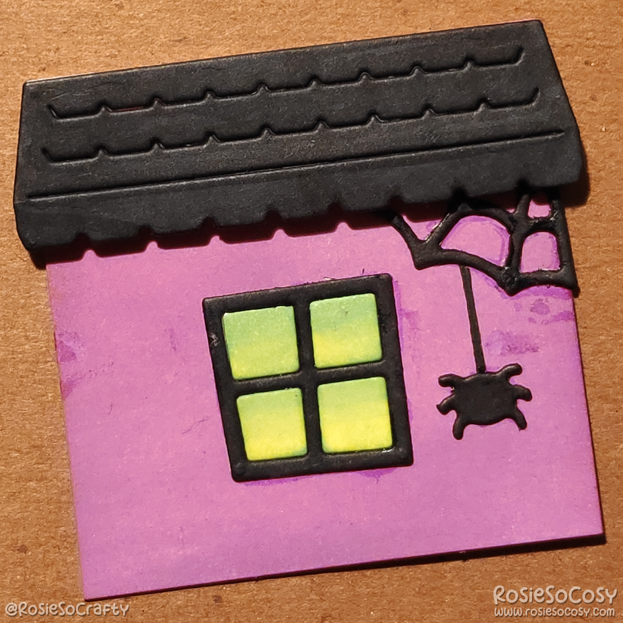 A two inch sized card consisting of a purple house, black window with green light coming from within the building, a black roof and a black spider hanging from a spiderweb in the top right corner.